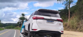 Guincho plataforma transportando um veículo branco, modelo SUV, em Mucuri, na beira da estrada.
