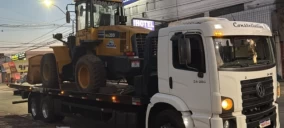 Guincho plataforma transportando uma retroescavadeira amarela em Abadia de Goiás, estacionado na rua.
