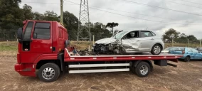 Guincho plataforma vermelho transportando um carro prata danificado em Videira, com vegetação ao fundo.