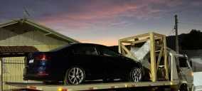 Guincho plataforma transportando um carro preto e uma estrutura de madeira em Rio do Sul ao entardecer.