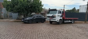 Guincho plataforma branco estacionado ao lado de um carro preto em uma rua de paralelepípedo em Carazinho.
